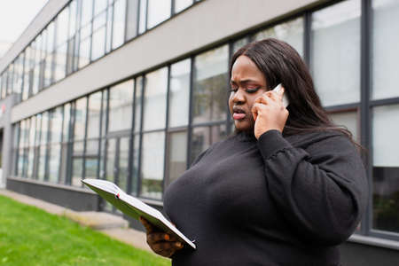 african american plus size woman holding notebook and talking on smartphone outsideの写真素材