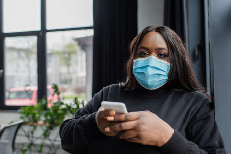 african american plus size businesswoman in medical mask using smartphone in office