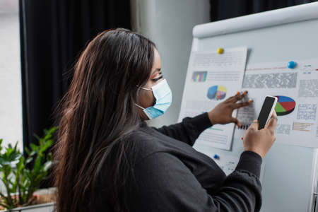 african american plus size businesswoman in medical mask using smartphone near flip chart with...