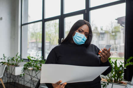 african american plus size businesswoman in medical mask using smartphone while holding documentの写真素材