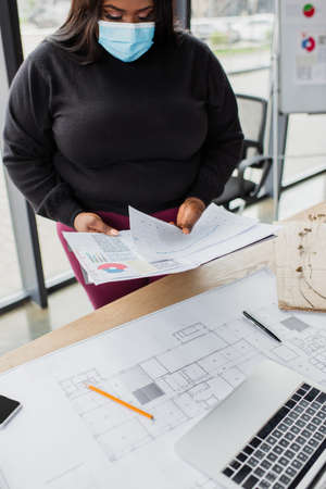 african american plus size engineer in medical mask holding charts and graphs near blueprintの写真素材