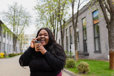 cheerful african american plus size woman holding coffee to go and talking on smartphone while laughing outsideの写真素材