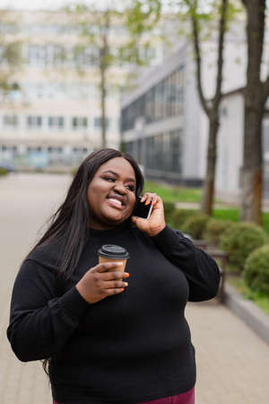 cheerful african american plus size woman holding coffee to go and talking on smartphone outsideの写真素材