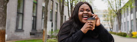 cheerful african american plus size woman holding coffee to go and talking on smartphone outside, bannerの写真素材