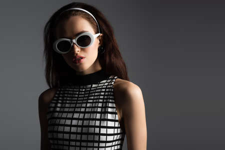 trendy woman in headband and white sunglasses looking at camera isolated on grayの写真素材