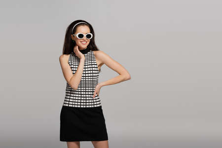 trendy young model in sunglasses, headband and retro outfit smiling isolated on grayの写真素材