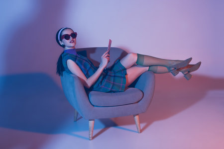 brunette young woman in sunglasses and retro outfit sitting in armchair with magazine on violetの写真素材