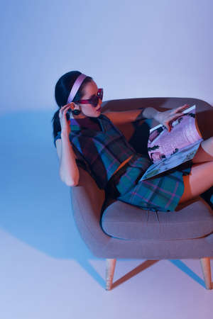 young woman in sunglasses and retro outfit sitting in armchair and reading magazine on blueの写真素材