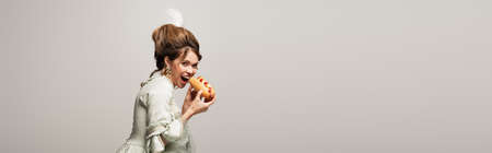 hungry, retro style woman looking at camera while eating hot dog isolated on gray, banner