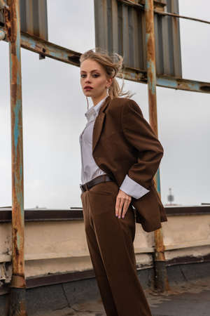 young model in earrings, white shirt and brown suit looking at camera while posing on rooftopの写真素材