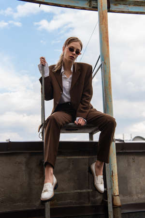full length of young model in stylish sunglasses and trendy suit sitting on metallic stairs on rooftopの写真素材