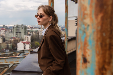 thoughtful woman in stylish sunglasses posing on rooftop with cityscape and blurred foregroundの写真素材
