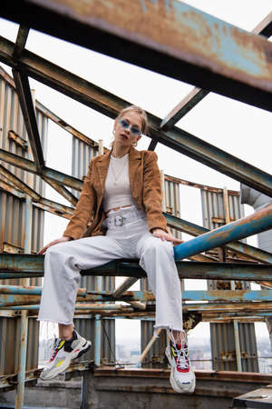 blonde woman in blue sunglasses and suede jacket posing on rusty construction of rooftopの写真素材