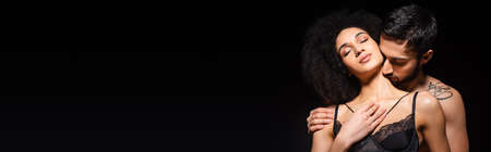 Young man kissing neck of african american woman in lace bra isolated on black, bannerの写真素材