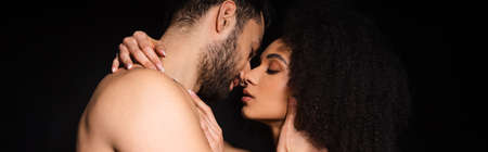 Side view of shirtless man kissing african american girlfriend isolated on black, bannerの写真素材