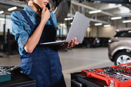 partial view of young african american mechanic holding hand near face and looking at laptop in...