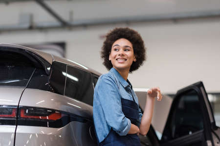 smiling young african american mechanic standing with near car in auto repair serviceの写真素材