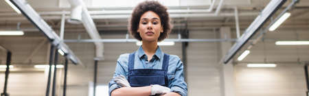 confident young african american mechanic standing with crossed arms and looking at camera in...