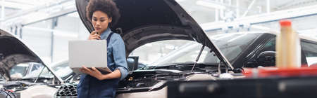 serious young african american mechanic holding laptop near car with open hood in auto repair service, bannerの写真素材