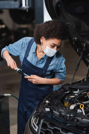 focused young african american mechanic in protective mask holding wrench and working with car motor...