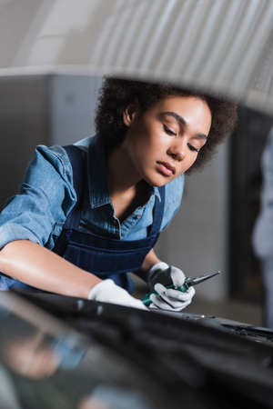 young african american mechanic holding pliers and fixing car motor in garage