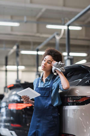 tired young african american mechanic standing with digital tablet and holding hand near head in...