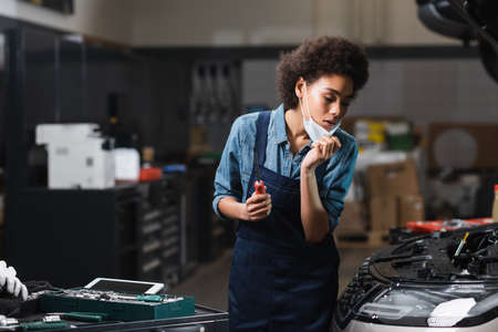 young african american mechanic holding protective mask with hand while working with motor of car in garageの写真素材