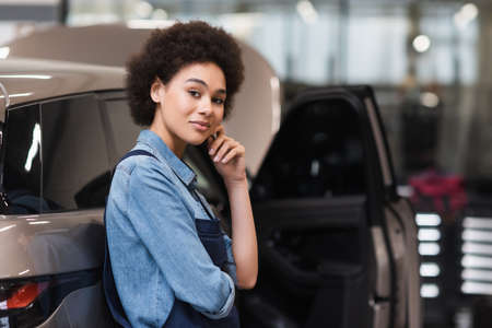 cheerful young african american mechanic standing with hand near face in auto repair service