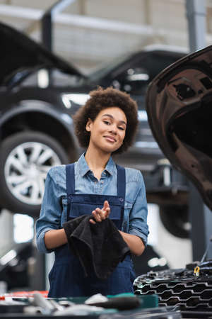 smiling young african american mechanic in overalls drying hands with black towel in garageの写真素材