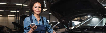 smiling young african american mechanic standing with cellphone near car with open hood in garage,...