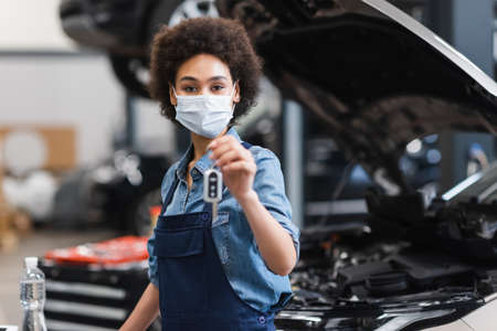 young african american mechanic in protective mask holding blurred car key in hand in garageの写真素材