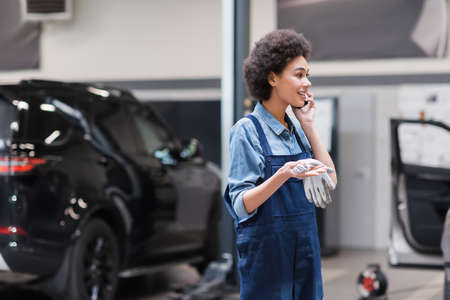 smiling young african american mechanic in overalls speaking on smartphone in auto repair serviceの写真素材