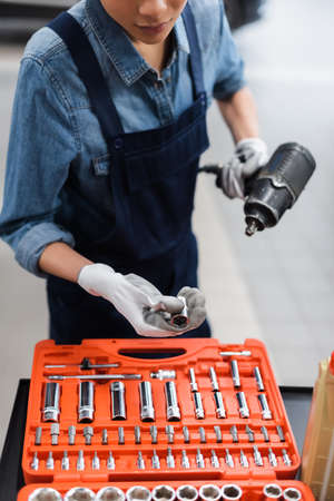 partial view of young mechanic with hands in gloves holding electric screwdriver near toolbox in auto repair serviceの写真素材