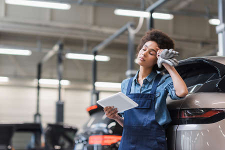 tired young african american mechanic with closed eyes standing with digital tablet and holding hand...
