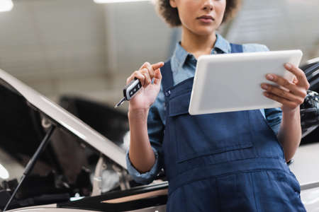 partial view of young african american mechanic in overalls holding car key and looking at digital tablet in garageの写真素材