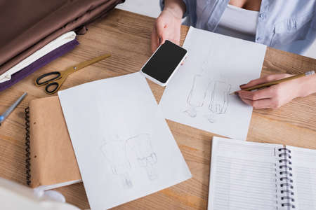 Cropped view of seamstress with smartphone drawing near cloth on table