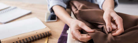 Cropped view of seamstress holding cloth near blurred cellphone and notebooks, bannerの写真素材