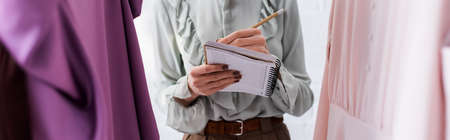 Cropped view of designer writing on notebook near clothes in atelier, bannerの写真素材