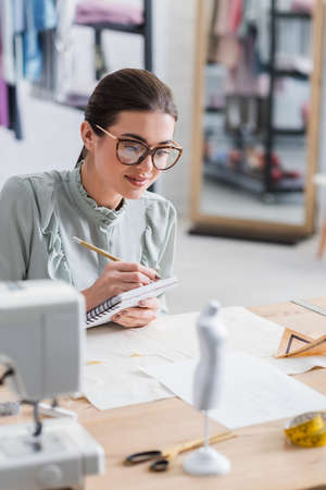 Smiling seamstress writing on notebook near mannequin and cloth on tableの写真素材