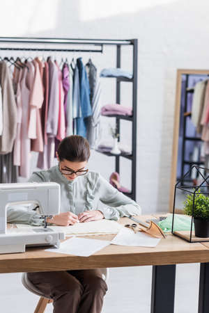 Designer marking fabric while working near sketches and sewing machine in studio