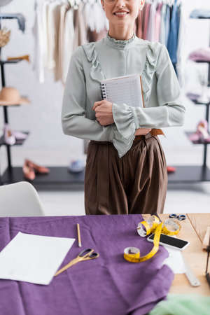Cropped view of smiling seamstress holding notebook near cloth, tape measure and cellphoneの写真素材