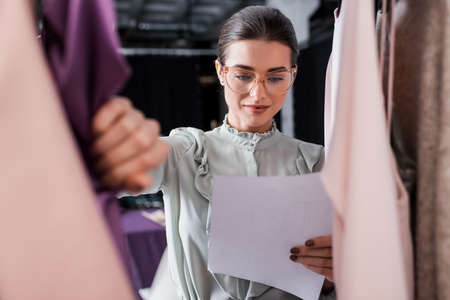 Seamstress in eyeglasses holding paper near blurred clothesの写真素材