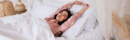 young asian woman in silk pajamas waking up with outstretched hands and closed eyes in bedroom,...