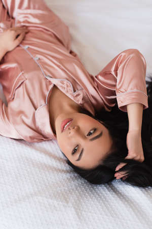 young asian woman in silk pajamas lying on bed and touching hair with hand in bedroom