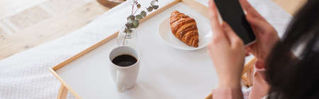 cropped view of young brunette woman holding cellphone near table with coffee and croissant in bedroom, bannerの写真素材