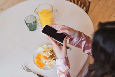 partial view of young woman holding cellphone and taking picture of pancakes with fruits near orange...