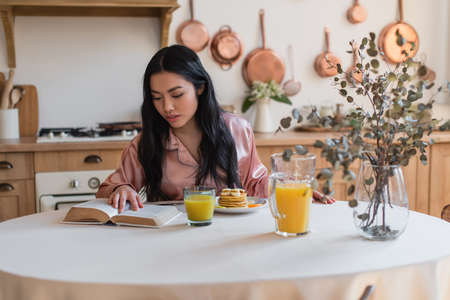 young asian girl in silk pajamas reading book while having breakfast in kitchenの写真素材