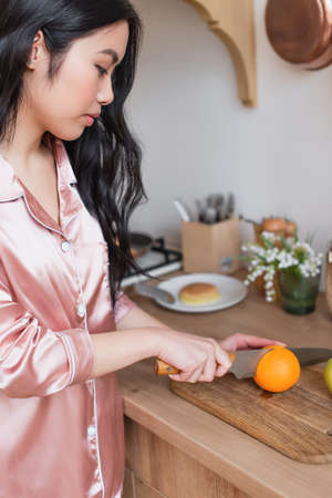 young asian woman in pink silk pajamas cutting orange in kitchen