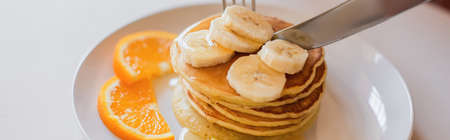 close up view of knife cutting pancakes with orange and banana slices on white plate, bannerの写真素材