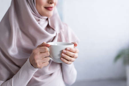 cropped view of blurred muslim woman in hijab holding cup of tea at homeの写真素材